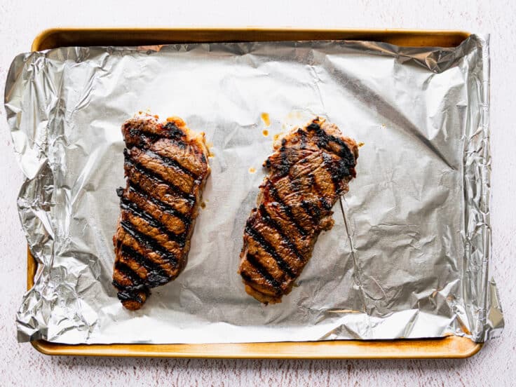 Grilled marinated New York strip steaks on a tray resting.