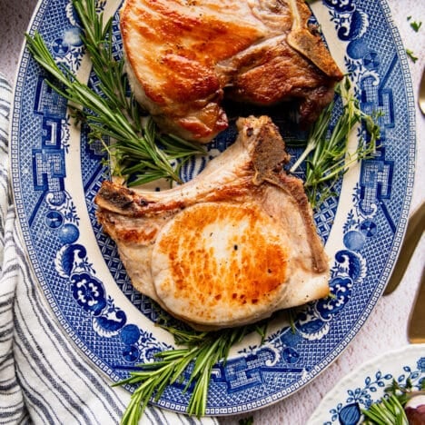 Square overhead shot of brined and seared pork chops on a blue and white platter with fresh herbs for garnish.