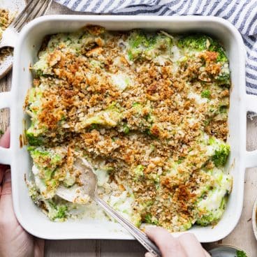 Square shot of a hand serving a broccoli casserole recipe from a white dish.