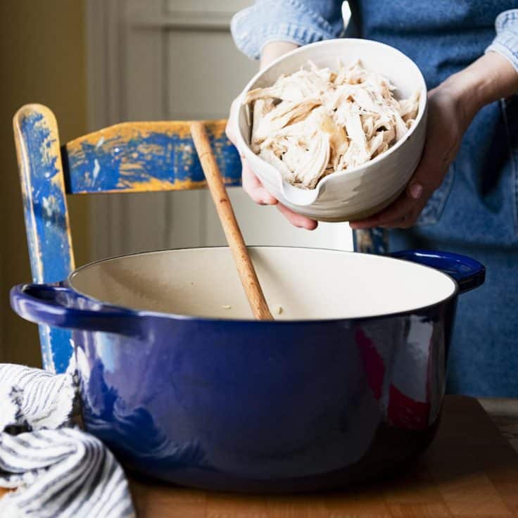 Adding shredded rotisserie chicken to a Dutch oven.