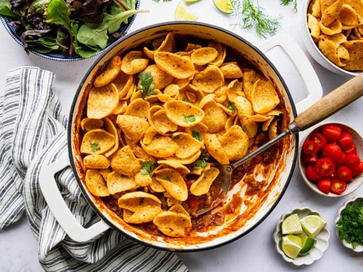 Horizontal overhead image of a Frito pie recipe on a white table with toppings and a side salad.
