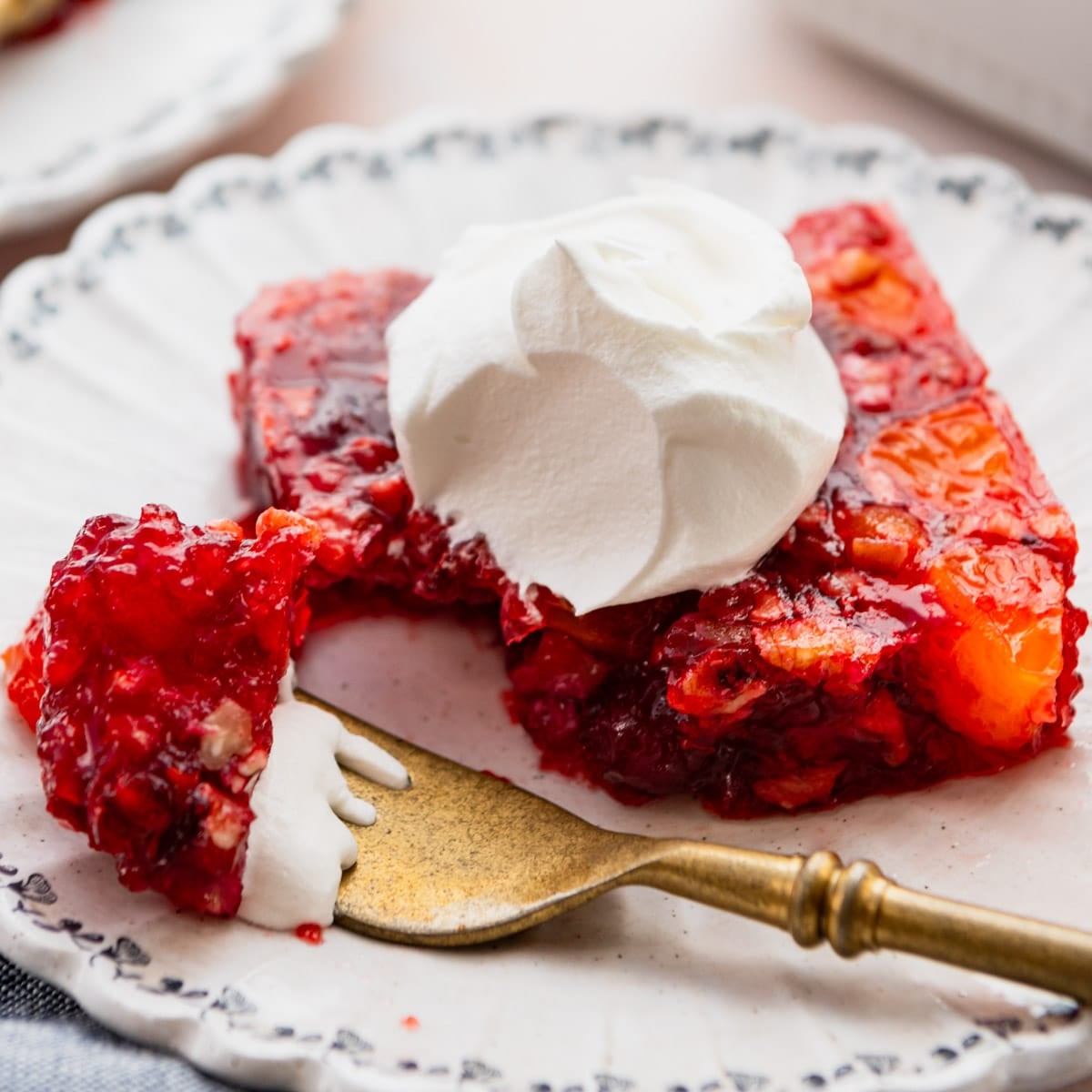 Square side shot of a square of cranberry jello salad on a small plate with a dollop of whipped cream on top.