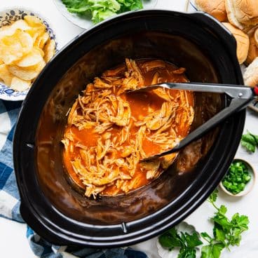 Square overhead shot of tongs in a pot of slow cooker buffalo chicken.