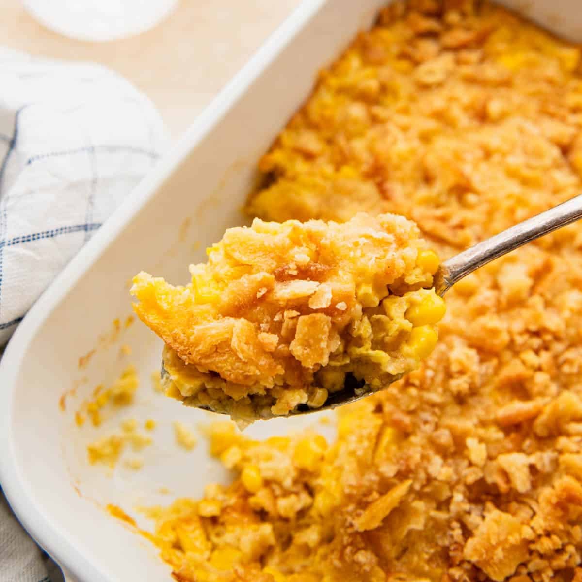 Serving spoon scooping scalloped corn from a baking dish to show the creamy texture and the crunchy topping.