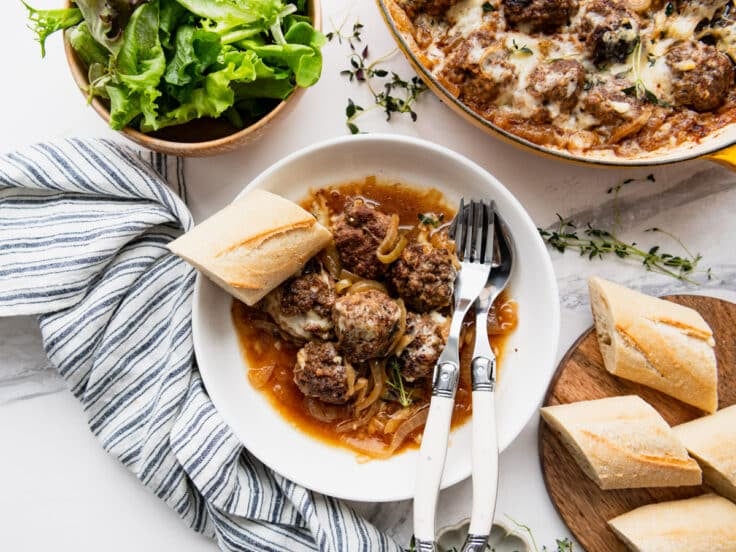 Horizontal overhead image of French onion meatballs in a white bowl with a baguette.
