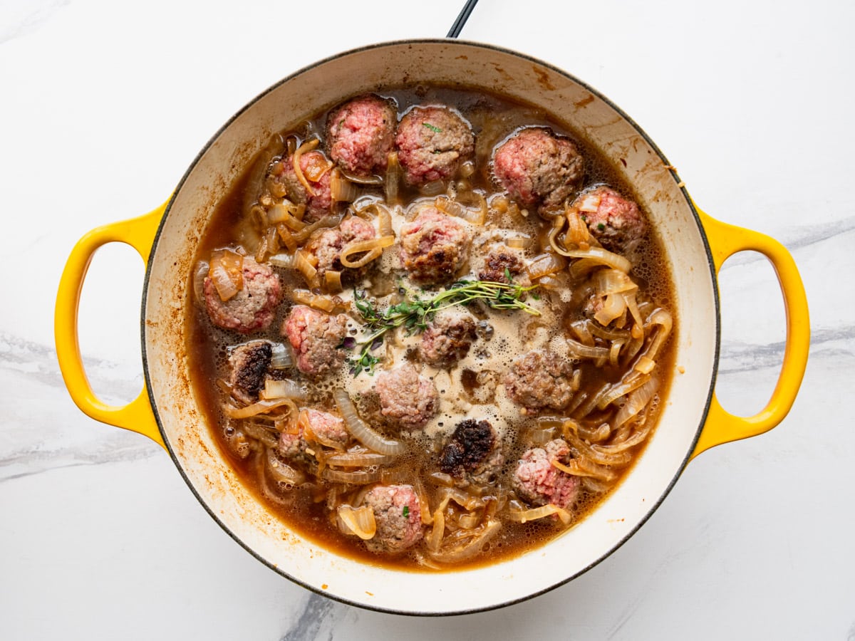 Adding broth, onions, and herbs to the skillet with the meatballs.