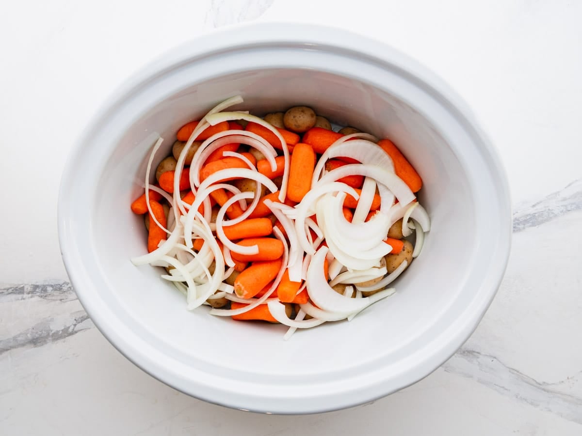 Potatoes and veggies in the bottom of a slow cooker.