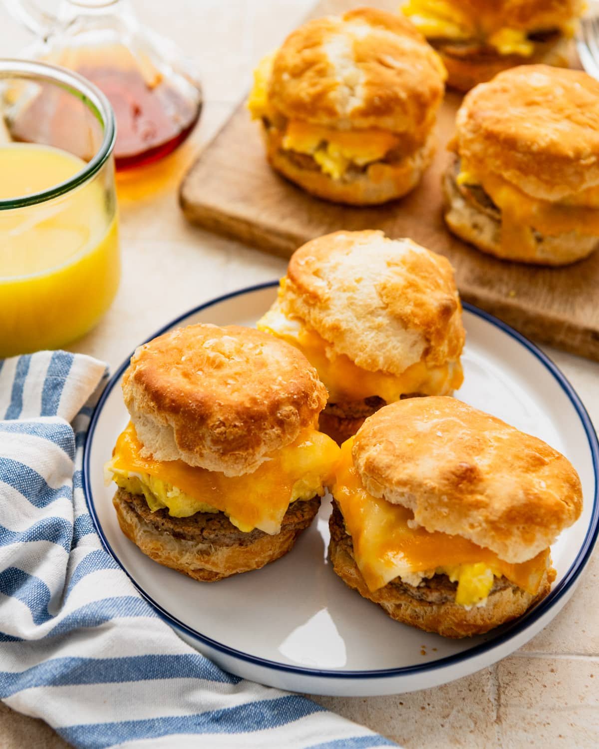 Side shot of breakfast biscuit sandwiches on a table with orange juice and syrup.