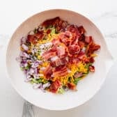 Combining the ingredients for a broccoli salad with bacon in a white mixing bowl.