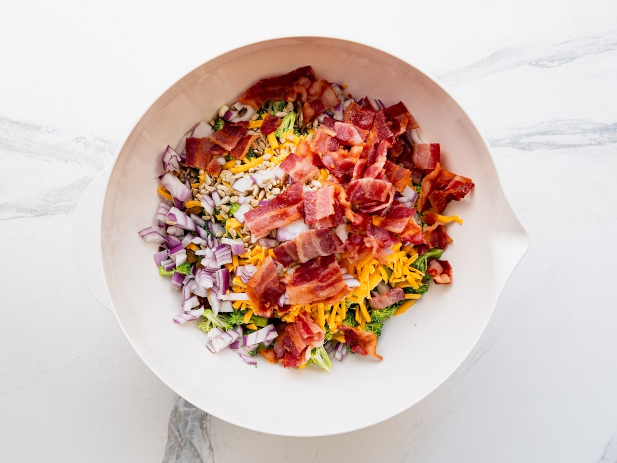 Combining the ingredients for a broccoli salad with bacon in a white mixing bowl.