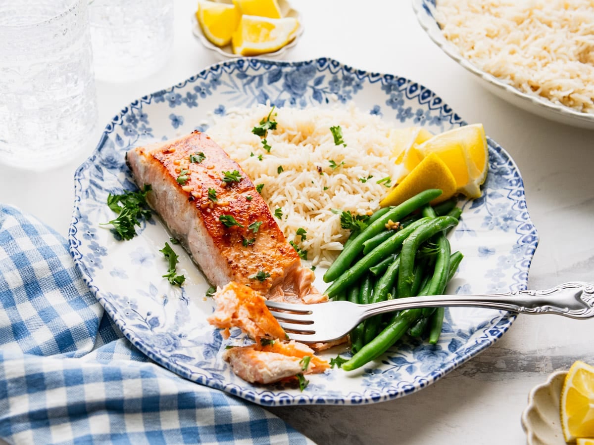 Horizontal side shot of a blue and white dinner plate full of garlic butter salmon, rice, and green beans.