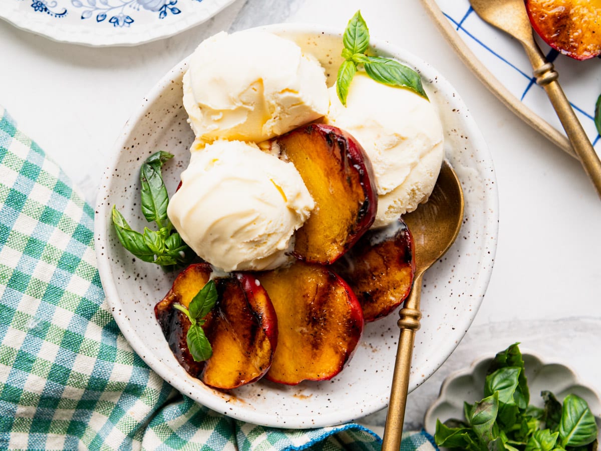 Horizontal overhead shot of a bowl of grilled peaches served with vanilla ice cream and garnished with fresh basil.