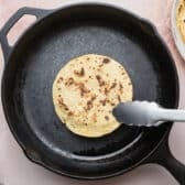 Using tongs to heat corn tortillas in a cast iron pan.