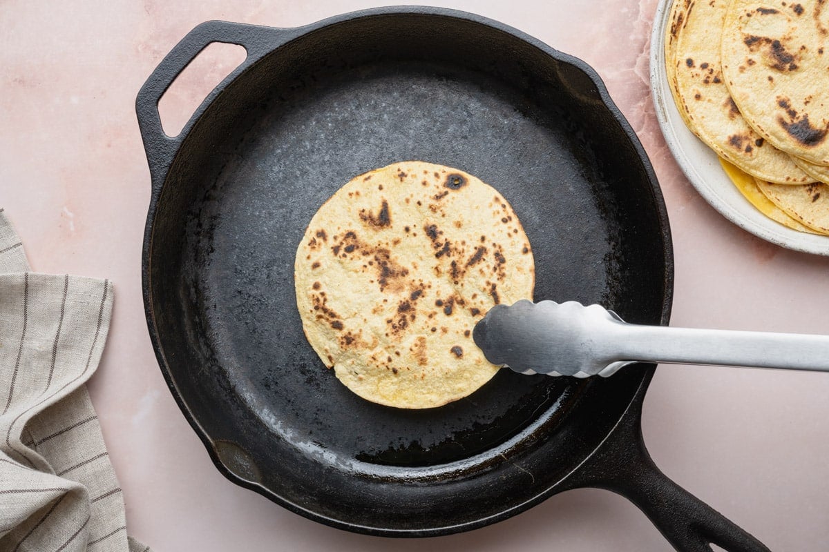 Using tongs to heat corn tortillas in a cast iron pan.