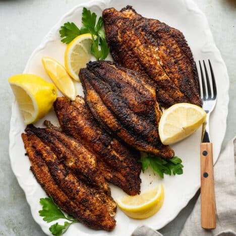 Square overhead shot of blackened catfish fillets on a white serving platter with fresh parsley and lemon wedges.