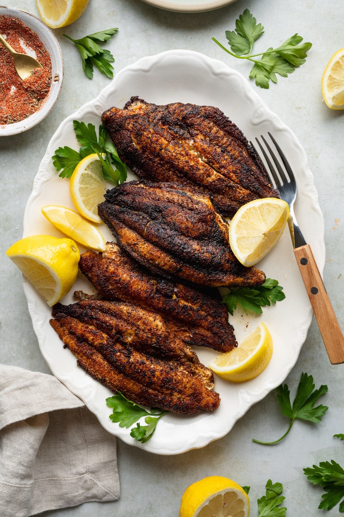 Overhead shot of blackened catfish fillets on a white serving platter with fresh parsley and lemon wedges.