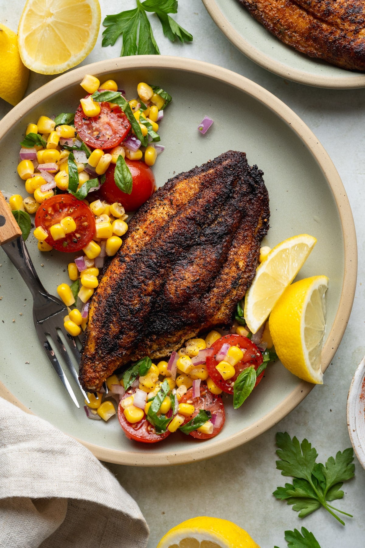 Overhead shot of blackened catfish on a plate with a side of lemon wedges and corn salad.
