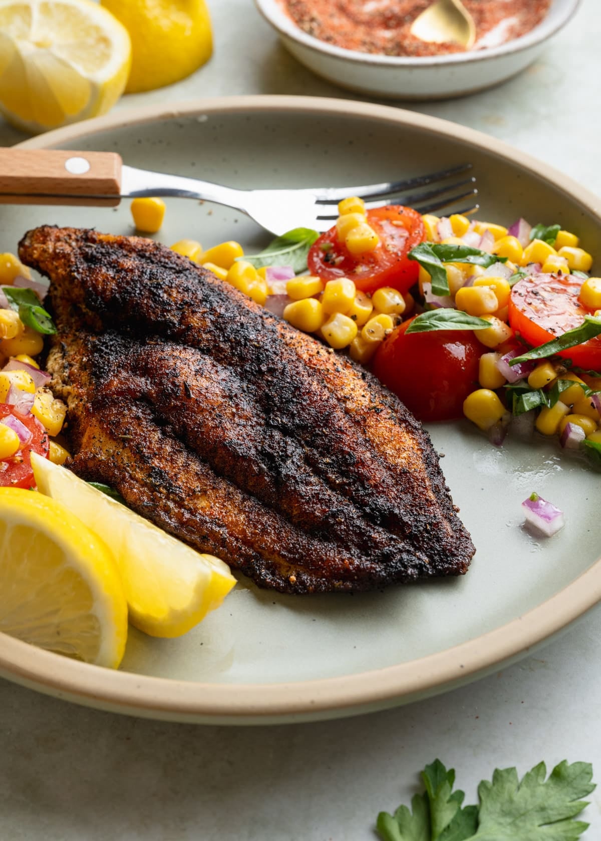 Side shot of blackened catfish on a plate with a side of lemon wedges and corn salad.