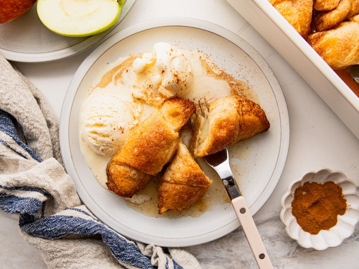 Horizontal overhead shot of two crescent roll apple dumplings on a white plate with a couple scoops of vanilla ice cream.