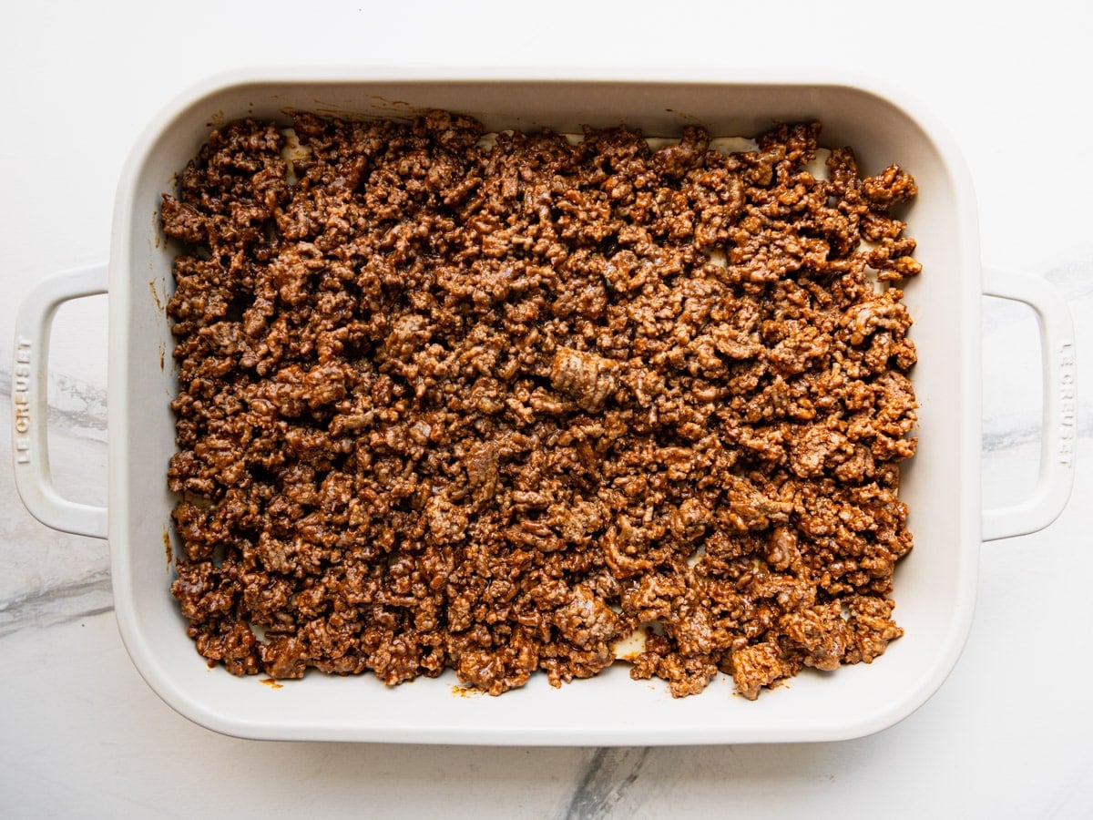 Spreading a layer of taco seasoned ground beef on top of the biscuit crust.