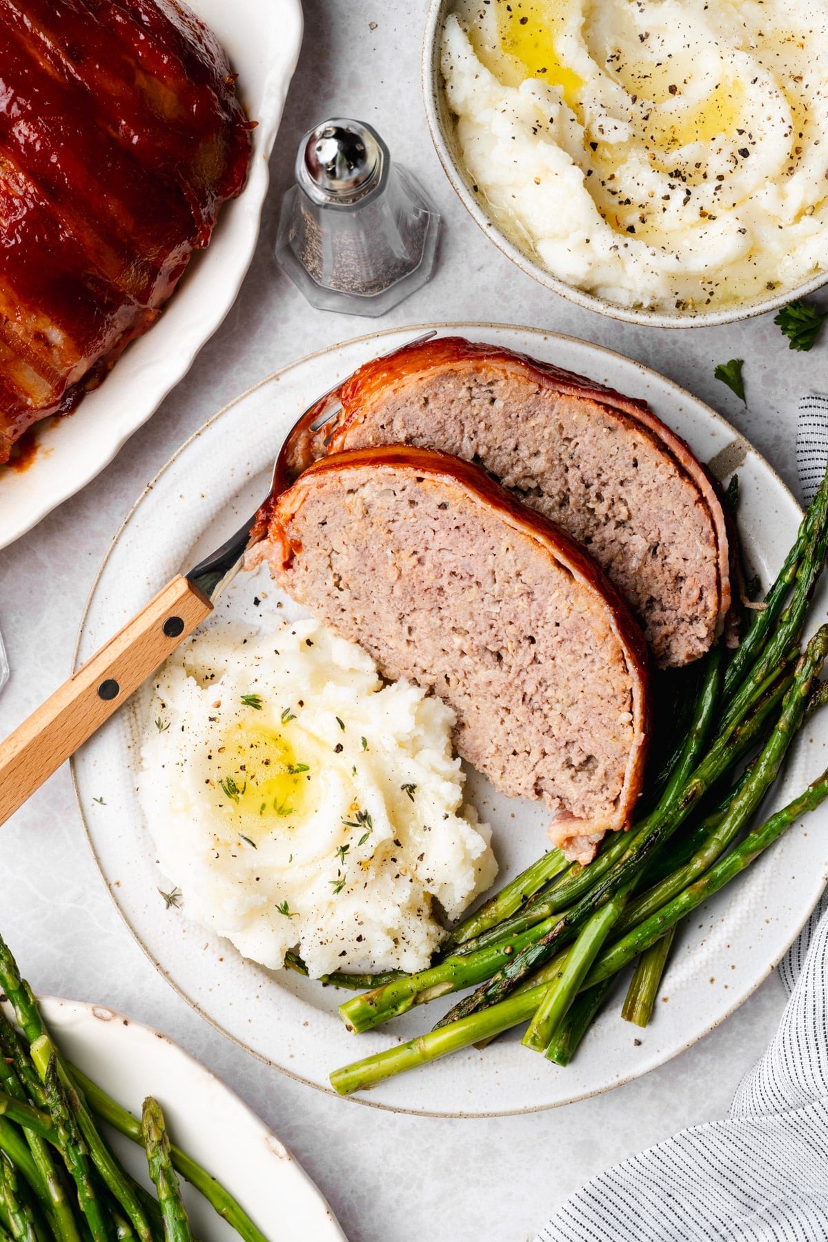 Overhead image of two slices of bacon wrapped meatloaf on a white plate with a side of mashed potatoes and asparagus.